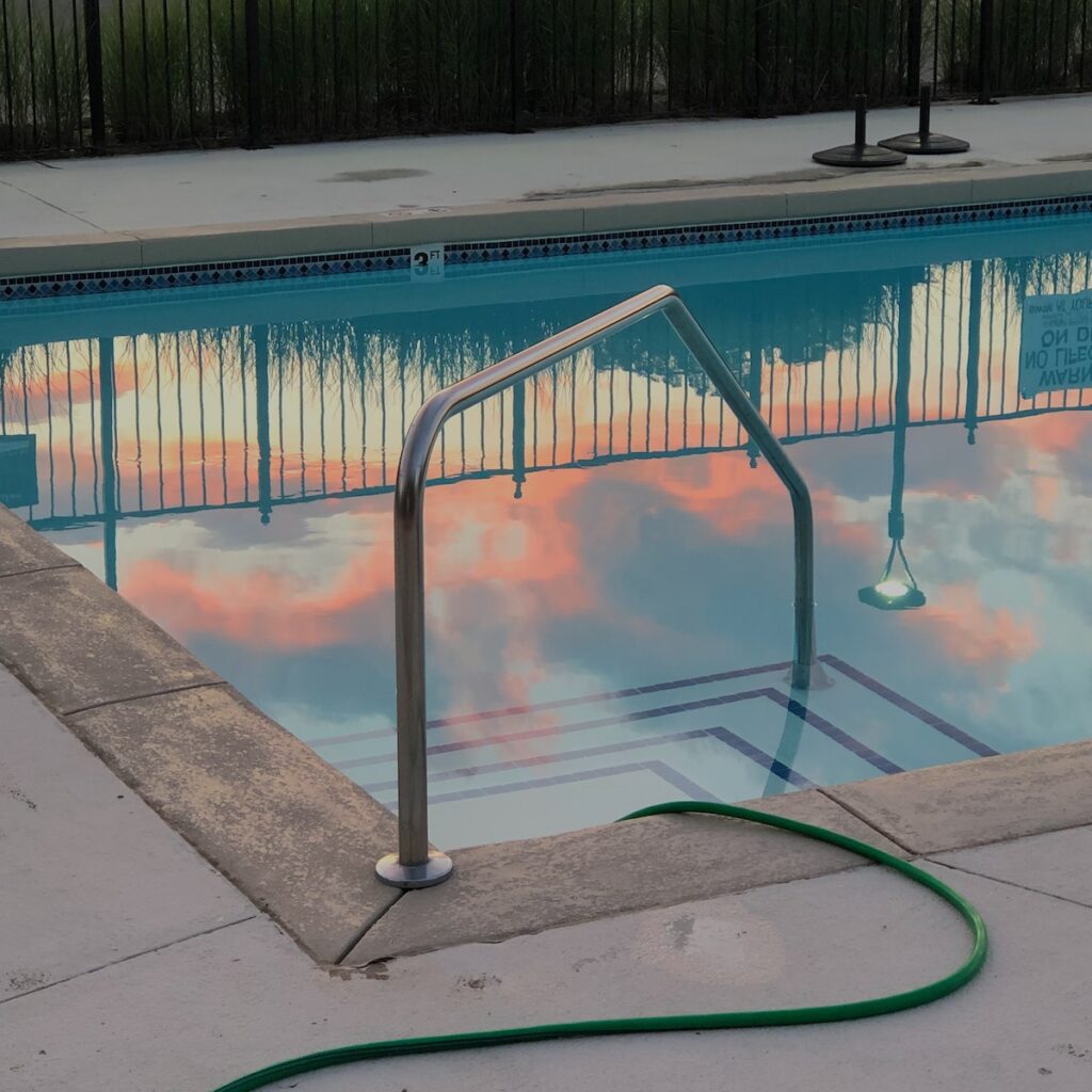 Empty small swimming pool with metal handrail and hose in backyard next to fence in evening